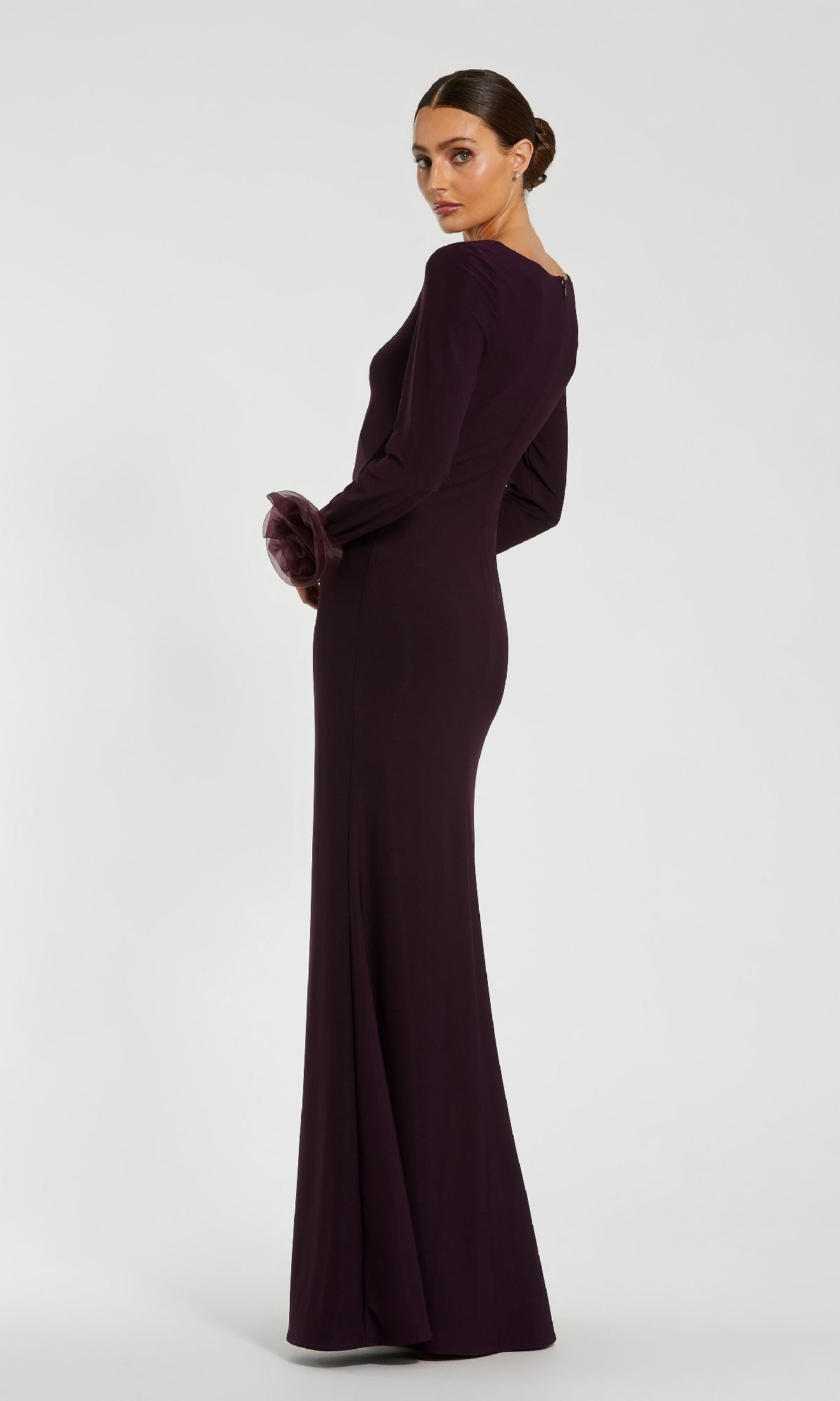 A woman stands against a plain background, looking over her shoulder in the Mac Duggal 49949 Long Sleeve Long Formal Dress—floor-length, fitted, and dark purple.